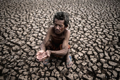 High angle view of shirtless man holding falling water while crouching on cracked land