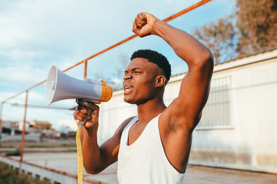 Young angry african american male in undershirt with speaker yelling with raised arm while looking at camera