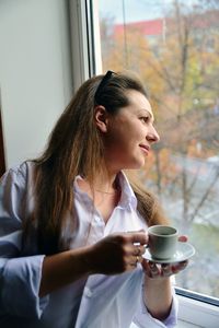 Young woman holding coffee cup while looking through window