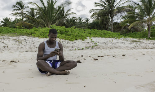 From the front, black traveler sitting on the sand and browsing social networks on cell phone