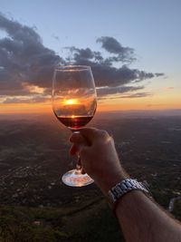 Man holding beer glass against sky during sunset