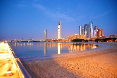 Illuminated cityscape by sea against sky during sunset