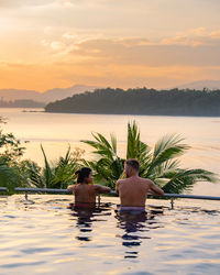 Rear view of shirtless man sitting in swimming pool against sea during sunset