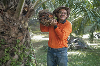 Portrait of man wearing hat holding palm fruit standing by tree