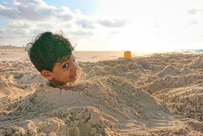 Portrait of boy buried in sand on beach against sky