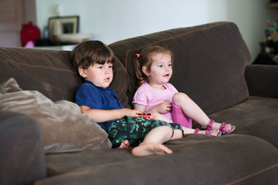 Siblings playing video game while sitting on sofa at home