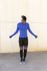 Young man exercising with jumping rope on footpath in front of wall