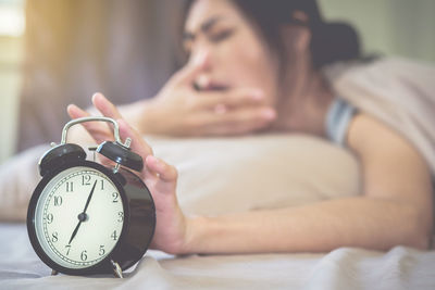 Woman yawning while holding alarm clock on bed