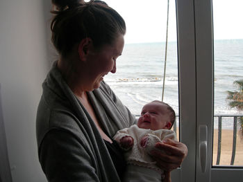 Side view of happy woman holding daughter by window at home