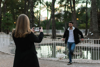 Man photographing woman using smart phone while standing on railing