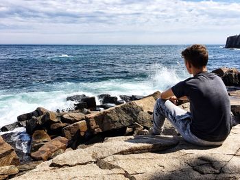 Rear view of man sitting on rock by sea against sky