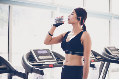 Young woman drinking water while standing in gym