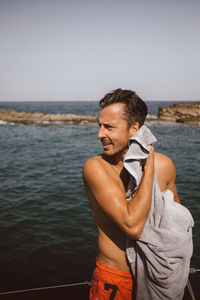 Man looking away while drying with towel at beach against sky on sunny day