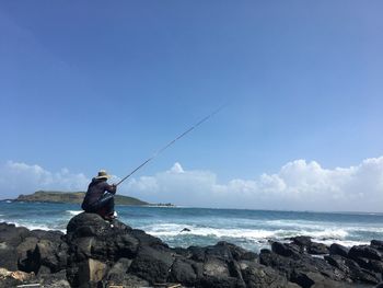 Man fishing on rock by sea against sky