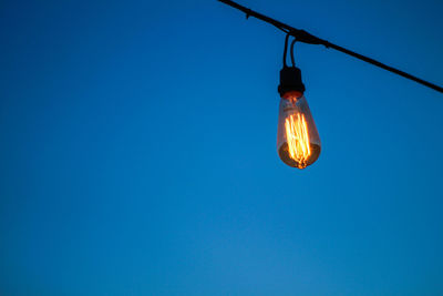 Low angle view of illuminated light bulb hanging against clear blue sky