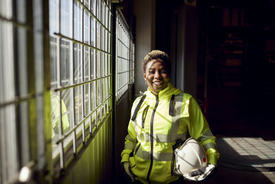 Portrait of smiling young female blue-collar worker standing by window in factory