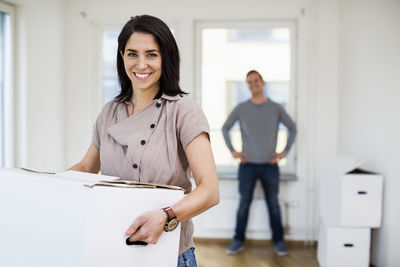 Portrait of happy woman carrying moving box with man in background at home