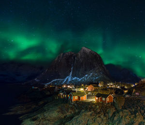 Illuminated houses by mountain against aurora borealis in sky at night