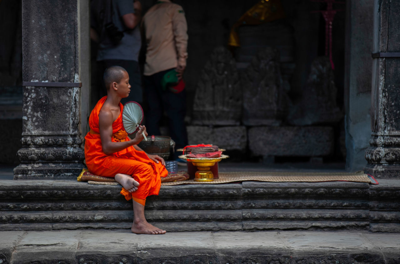 full length, architecture, real people, one person, built structure, day, building, men, staircase, building exterior, traditional clothing, religion, sitting, clothing, spirituality, orange color, lifestyles, outdoors, child