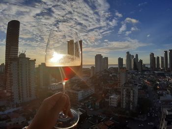 Man holding glass of buildings in city against sky