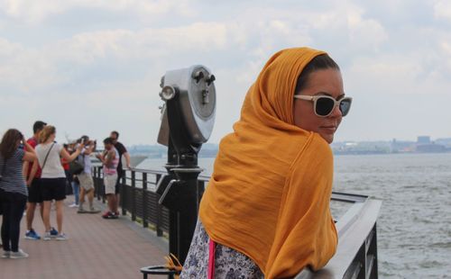 Woman with headscarf and sunglasses leaning on railing at liberty island against sky