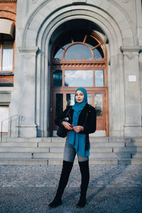 Full length portrait of confident young muslim woman standing with smart phone against entrance door