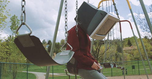 Man wearing television set while sitting on swing at playground