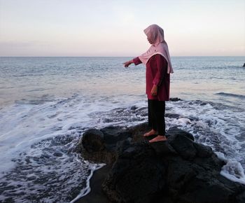 Full length of woman standing on rock at beach against sky