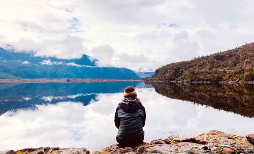 Rear view of woman sitting by lake against cloudy sky during winter