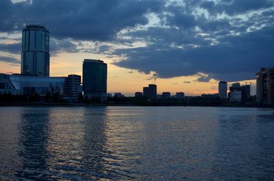 Modern buildings by river against sky during sunset