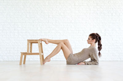Full length of young woman leaning while pushing stool against brick wall