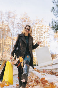 Woman kicking autumn leaves walking on footpath holding shopping bags