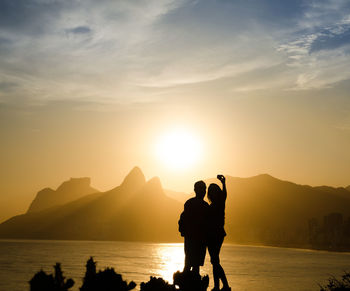 Silhouette couple taking selfie by sea against sky during sunset