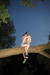 Couple kissing while sitting on tree against sky
