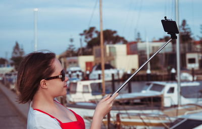 Side view of young woman holding monopod while taking selfie through smart phone at harbor