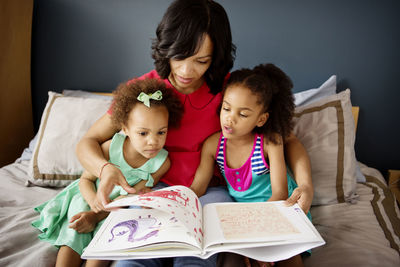 Mother and children looking in book while sitting on bed at home