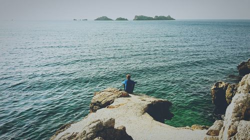 Rear view of man sitting on rock formation in sea against sky