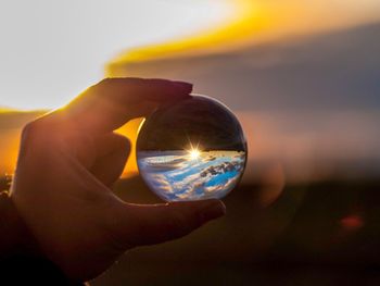 Close-up of hand holding crystal ball against sky during sunset