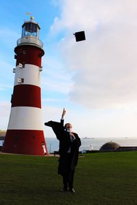 Student throwing mortarboard while standing against lighthouse