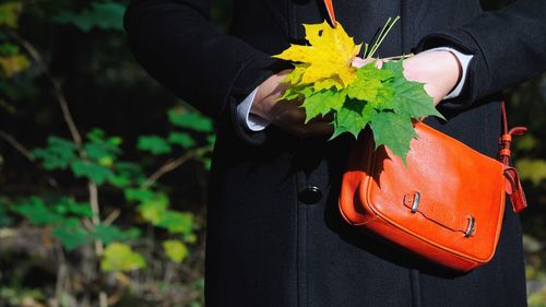 Midsection of woman standing with maple leaves with purse on field