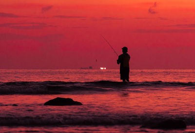 Silhouette man fishing in sea against orange sky