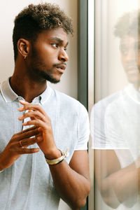 Serious young black bearded male with curly hair in casual shirt looking away thoughtfully while standing near window