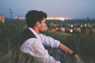 Side view of young man looking away while sitting on field against sky during sunset