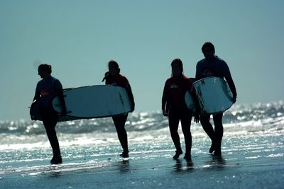 Silhouette surfers carrying surfboards on shore against clear sky