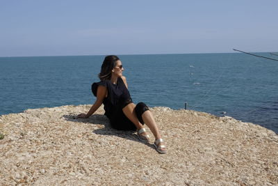 Young woman sitting on rock at beach against clear sky