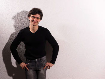 Portrait of smiling young man standing with hands in pockets by white wall