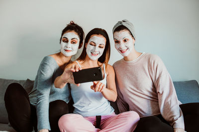Cheerful female friends with facial masks taking selfie against wall at home