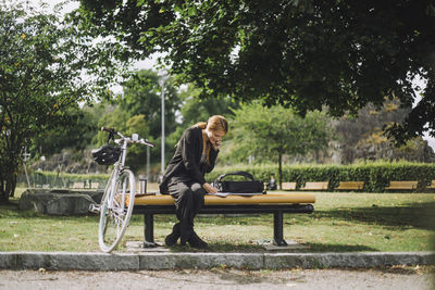 Full length of businesswoman talking on smart phone while sitting by bicycle at park