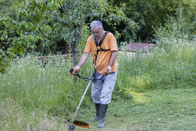 A mature man in protective clothing, and gloves  and mowing tall grass and the weeds with a trimmer