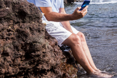 Low section of man with smart phone sitting on rock by sea